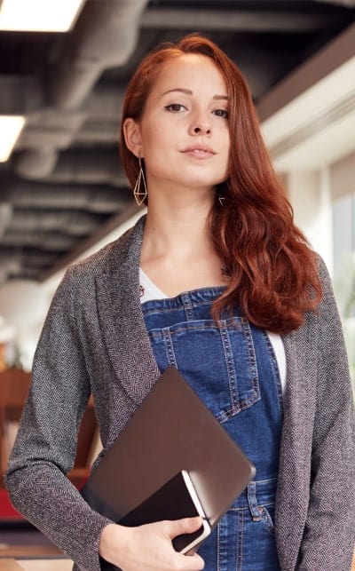 Vrouw met lang rood haar en blazer, holding laptop in kantoor.